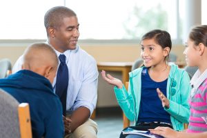 Diverse group of elementary age little girls and little boys are sitting in circle with mid adult African American male counselor. 